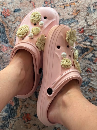 A pair of light pink Crocs, decorated with cream-colored flower charms and faux pearls, are worn on a person's feet. The Crocs are positioned on a patterned rug with floral designs in shades of red, blue, and orange. The scene is shot from a high angle, looking down at the feet and shoes. The environment does not provide any specific indicators of location within Kumbor, Montenegro, or any other city.