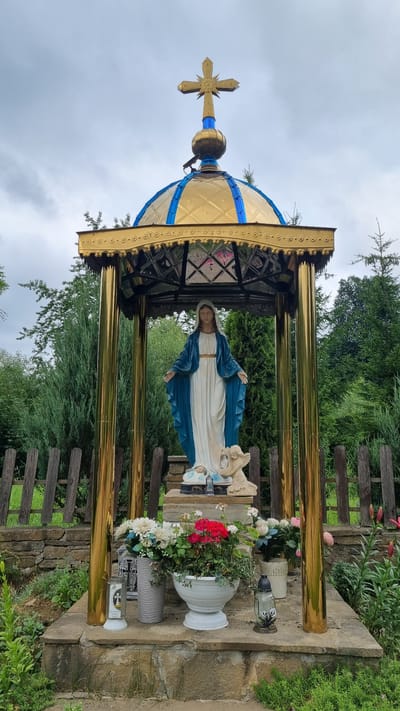 The media file depicts a religious shrine with a statue of the Virgin Mary. The shrine is adorned with a golden dome topped with a cross, and supported by golden pillars. The statue of the Virgin Mary, dressed in a blue mantle and white robe, stands with her arms outstretched. She is accompanied by a smaller figure, possibly representing an angel or child.
The shrine is situated outdoors, amidst lush greenery and trees. A wooden fence runs along the background. In front of the shrine, various offerings are placed, including a large white urn filled with red and white flowers, another vase with white flowers, and a candle holder with a lit candle, featuring Christian symbols. The ground appears to be made of stone. The overall scene suggests a place of worship or remembrance, possibly a roadside shrine or a memorial site.
The lighting suggests it is daytime, and the overcast sky indicates possibly cloudy weather. The location is identified as Vyshka, Ukraine. There are no visible signs of any specific event taking place, other than the presence of religious offerings, which implies ongoing devotion. The mood conveyed is one of reverence and peace. The statue itself shows no visible text, nor does the immediate surroundings, apart from the Christian symbols on the candle holder.