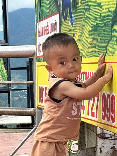 In the city of Tu Le, Vietnam, a young East Asian child with short, dark hair is positioned in the foreground, facing slightly right of the viewer with an inward gaze. The child wears a light brown sleeveless top displaying "STA" and "The greate" (partial) in white text, over a dark undergarment, and matching shorts. A thin, light-colored bracelet is on the child's right wrist, and a dark strap is around the neck. Both of the child's hands are pressed against a large, vertical sign that is predominantly yellow with a green upper section. The green section depicts a pattern resembling terraced fields and a blue human figure's legs. The sign features Vietnamese text in white on a red horizontal bar: "NHAN NHIEU Uu DAI," and red numbers on the yellow background, including "0915" (partial) on the left and "727 999" on the right. The sign is mounted on weathered metal posts. In the mid-ground to the left, a rusted metal railing runs horizontally. Behind the railing and the sign, hazy blue mountains and distant green foliage are visible, indicating an elevated outdoor setting. The ground directly beneath the child is a reddish-brown paved surface.