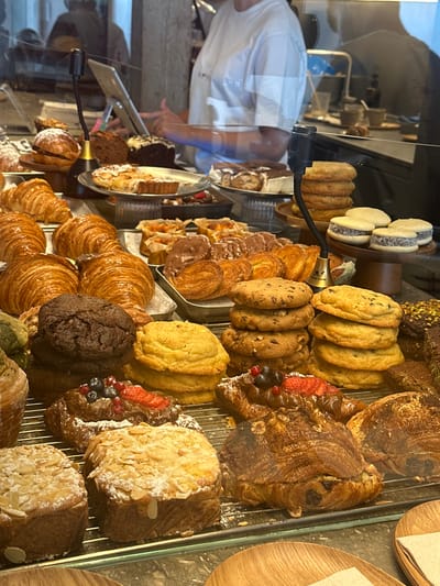 The image depicts the interior of a bakery or cafe in Santander, Spain, specifically focusing on a glass display case filled with a variety of baked goods. In the background, visible through the reflective glass, a person in a white short-sleeved t-shirt operates a tablet on a stand, positioned on a counter.
The foreground is dominated by multiple tiers of pastries and desserts arranged on metal racks, plates, and wooden stands. From top to bottom and left to right, visible items include:
* Several stacks of golden-brown croissants on white trays.
* A dark rectangular cake with white frosting, and a round fruit tart on a silver stand.
* Another rectangular cake, possibly a cheesecake, on a silver stand.
* Stacks of round cookies (some chocolate chip, some plain) on wooden stands.
* Three white, round alfajores with visible dulce de leche filling on a wooden stand.
* Various smaller pastries including palmiers, glazed pastries, and muffin-like items.
* On the lowest tier, a prominent dark chocolate muffin or large cookie.
* Stacks of light-colored, round cookies.
* Square pastries topped with fresh red currants and blueberries.
* Square pastries topped with sliced almonds and powdered sugar.
* Large, textured, layered pastries, some with visible nuts.
A flexible black stand with a lamp or microphone head, labeled "REALTIME," is positioned in the middle of the display, slightly behind the front items. The overall environment suggests a commercial setting designed for food service.