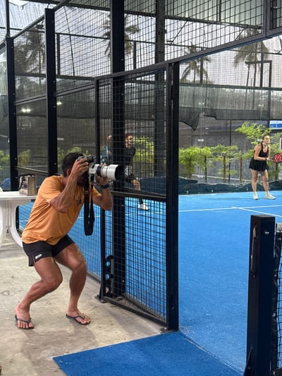 In Baan Plai Laem, Thailand, a photographer wearing an orange t-shirt and black shorts crouches behind a black metal fence, holding a large camera with a telephoto lens to his eye. He is focused on a woman playing padel on a bright blue court. The woman, wearing a black sleeveless dress and white sneakers, is holding a padel racket and appears to be in the middle of a game. The court is enclosed by black metal fencing with mesh panels, and palm trees are visible in the background, suggesting a tropical setting. The ground outside the court is a concrete-like material. A white table with a bag on it is visible to the left of the photographer.
