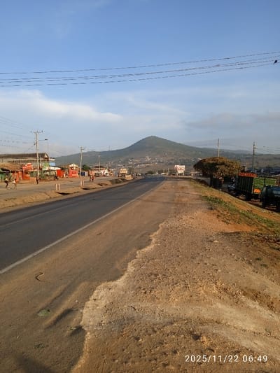 An empty, straight two-lane road stretches towards a distant, rounded mountain under a blue sky with scattered clouds. Buildings and roadside stalls line the left, while parked trucks and vegetation occupy the right. Utility poles with wires run parallel to the road. The foreground features a wide, eroded shoulder transitioning into a dirt embankment. A timestamp is overlaid in the bottom right corner.