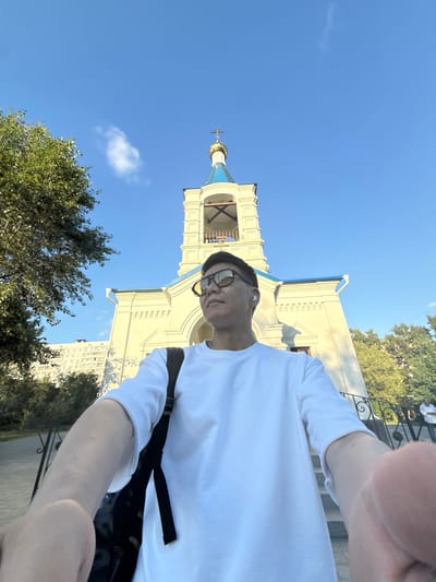 The image is a low-angle, selfie-style shot taken outdoors on a sunny day, with a clear blue sky. In the foreground, a person wearing a white t-shirt, sunglasses, and white wireless earbuds is visible, holding the camera out to capture the scene. A black backpack is slung over their left shoulder. The person is looking upwards and to the right.
Behind the person, a light-colored building with a blue roof and a golden dome topped with a cross is prominently featured. This appears to be a church or a similar religious building. The architecture is classical, with arched windows and decorative elements. To the left of the building, a large green tree with dense foliage is visible. In the background, a tall apartment building with numerous windows can be seen. A low black metal fence with ornate scrollwork runs along the lower part of the frame, separating the viewer from a paved walkway or path.
The specific location is identified as the Church of the Nativity of the Blessed Virgin Mary in Dolgoprudny, Russia. The building's architecture, including the dome and cross, aligns with common designs of Russian Orthodox churches. The presence of a modern apartment building in the background suggests an urban or suburban setting, typical for many churches within Russian cities. The overall atmosphere is bright and clear, indicative of a pleasant daytime.