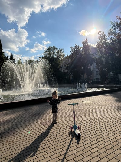 This media file captures a sunny day in Voronezh, Russia, likely during the summer. The scene is a public park or square featuring a large fountain and a paved walkway.

The main subjects include a young girl and a scooter. The girl, wearing a dark dress with a floral pattern, is standing near the edge of the fountain, looking down at it. Her posture suggests she might be interacting with or observing the water. A child's scooter, with a light-colored deck and pink handlebars, lies on the paved walkway to the right of the fountain.

The fountain is active, with multiple jets of water spraying upwards and outwards, creating a shimmering effect in the sunlight. The walkway is paved with rectangular bricks arranged in a herringbone pattern. In the background, trees and buildings are visible, suggesting an urban environment. The sky is bright blue with scattered white clouds, and the sun is prominently visible, casting strong shadows and lens flares, indicating it is likely mid-day.

Notable details include the strong sunlight and the lens flare emanating from the sun, suggesting a clear and bright day. The presence of trees and buildings provides context for an outdoor public space. The overall mood appears to be one of a pleasant, leisurely day outdoors. There is no visible text in the image.