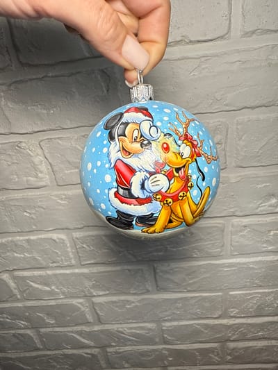 A person's hand holds a Christmas ornament against a grey brick wall. The ornament is round and blue with white polka dots. It features a drawing of Mickey Mouse dressed as Santa Claus hugging Pluto, who is dressed as a reindeer with antlers and bells. Mickey is smiling and has a white beard and fluffy trim on his Santa suit. Pluto has a red ribbon tied around his antlers and a red collar with bells. The background appears to be indoors, and no specific landmarks of Lipetsk, Russia, are visible.