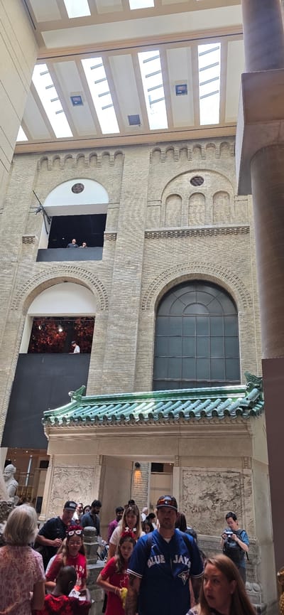 The image shows a group of people standing and looking towards a building. The building appears to be made of brick and has several windows. The people are dressed casually, and some are wearing baseball caps. The scene is outdoors, and the lighting suggests it is daytime. The people appear to be on a tour or visiting a historical site. The overall mood seems relaxed and observational.
One person is wearing a blue Toronto Blue Jays jersey with "JAYS" and "BLUE" visible on the back. Another person is wearing a red shirt. There is a large, ornate green structure on the right side of the image. The background includes architectural details such as arched windows and decorative brickwork. The individuals are looking upwards and towards the building's facade. There are no clear indicators of the time of day or weather conditions beyond the general daylight. The location cues suggest an urban setting, possibly a museum or public building in Toronto, Canada, given the Blue Jays jersey.