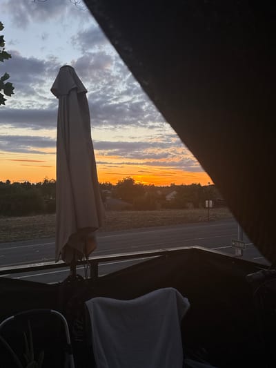The image captures a sunset view from a covered outdoor area, possibly a patio or balcony, in Sacramento, California. The sky is filled with a dramatic mix of orange and pink hues near the horizon, transitioning to grays and blues overhead. Scattered clouds add texture to the sky. In the foreground, a closed umbrella stands tall, draped with a fabric cover. To the right, a dark, amorphous shape obscures part of the scene, suggesting it is the edge of the covered area.

Beyond the immediate foreground, a road runs horizontally across the image, bordered by dry, grassy fields. In the middle ground, a line of trees and houses forms the silhouette of a suburban landscape. A street sign with an arrow pointing to the left and the word "LIBRARY" is visible on the right side of the road, indicating the presence of a library in that direction.

The lighting suggests it is either sunrise or sunset, with the warm tones of the sky dominating the scene. The overall atmosphere is tranquil and evokes a sense of dusk. There are no people or animals clearly visible in the image. The scene appears to be in a residential or semi-rural part of Sacramento, given the houses and road.