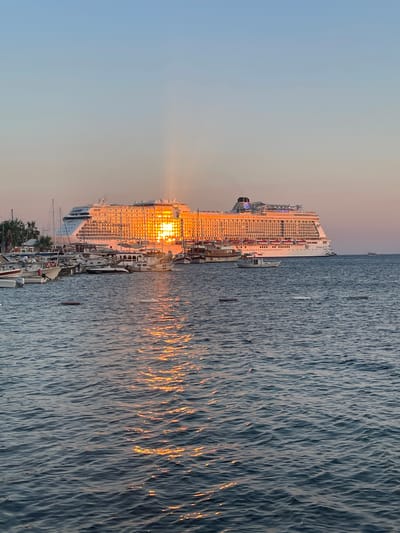 The image captures a serene sunset at a harbor in Bodrum, Turkey. The primary subjects are a large cruise ship docked alongside several smaller boats and yachts, all bathed in the warm, golden glow of the setting sun. The scene is set on the water, with the calm sea reflecting the fiery hues of the sunset.
The cruise ship, a prominent object in the frame, is illuminated by the sun, with its windows and decks shimmering. Smaller vessels are moored alongside, creating a picturesque harbor view. The activity depicted is a peaceful evening scene at a port, with the ships docked and the sun setting over the horizon.
Notable details include the time of day, clearly indicated by the sunset, which casts long rays of light and creates a warm, inviting atmosphere. The water is rippled, with the sunset's reflection creating a vibrant, textured pattern. The sky transitions from a soft blue to a warm orange and pink hue near the horizon, typical of a beautiful sunset. Although no people are explicitly visible, the presence of the docked ships suggests a bustling port that is winding down for the evening. The location is identified as Bodrum, Turkey, which contributes to the Mediterranean ambiance of the scene. The overall mood evoked is one of tranquility and natural beauty. There is no visible text in the image.