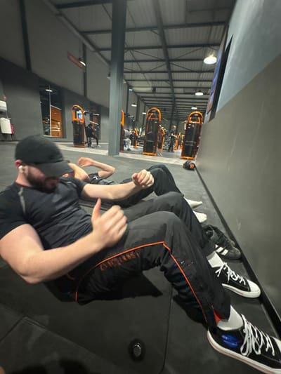A side-angle shot within a gym shows a man with a beard, wearing a black cap and shirt, engaged in a sit-up exercise. He is positioned on a black mat. Behind him, a line of people are similarly exercising on mats. Gym equipment is visible in the background, arranged in rows under bright overhead lighting. The man's legs are positioned near the top of the frame, showcasing white socks and dark sneakers.
