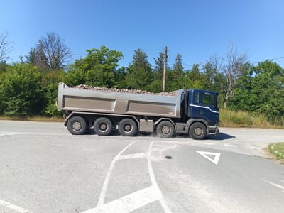 A dark blue dump truck, loaded with rocks, is captured in a side-view profile on a paved road intersection under a clear blue sky. The truck is in motion, indicated by dust kicked up by its rear wheels. The road surface has white directional markings. The truck is positioned on the right side of the frame, with its front facing right. The background consists of dense green trees and vegetation. The scene appears to be in Golyam Manastir, Bulgaria, given the context.