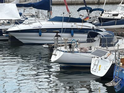 The image depicts a harbor in Wismar, Germany, with several sailboats moored. In the foreground, a white sailboat with blue trim is prominently featured, its name "JacSa" visible on its hull. This boat has a blue fender hanging on its side and a small motor attached to the stern. Behind it, another sailboat with a blue canopy is partially visible, and further back, more boats and masts are present. The water is choppy, reflecting the boats and the overcast sky. The scene appears to be a typical view of a marina, with the boats indicating recreational or possibly professional maritime activity.