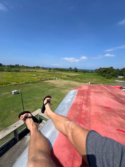 This image captures a person's legs and feet, clad in dark flip-flops, as they are positioned at the edge of a red structure, possibly an aircraft wing or a platform. The person is wearing grey shorts. The perspective is from a high vantage point, looking down and to the side.

Below the elevated structure, a vast expanse of green, grassy terrain stretches out, dotted with patches of vegetation and some agricultural fields. In the distance, a line of trees forms a dense border, beyond which a glimpse of the ocean is visible under a clear blue sky with scattered white clouds. The bright sunlight suggests it is daytime, likely mid-morning or afternoon, and the weather appears fair.

The scene evokes a sense of being in an open, possibly rural or coastal area in Blahbatuh, Indonesia. The elevated position and the vast landscape create a feeling of openness and perhaps a touch of adventurousness or contemplation. There are no visible text elements in the image.