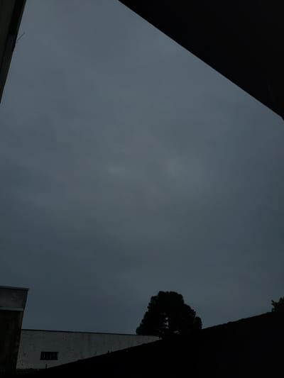 The image offers an upward perspective of a heavily overcast, uniformly grey sky, framed by dark, silhouetted structures on the left, right, and bottom edges. The central focus is the expansive, muted grey sky, indicating dense cloud cover during the daytime.

On the right side of the frame, a tall, light-colored building with a textured, possibly weathered, surface rises vertically. A small, dark, rectangular window opening is visible on its upper facade. Adjacent to this building, the dark, dense silhouette of a tree canopy is prominent against the sky, partially obscuring the building. The structures on the left and bottom are deep black, providing a stark contrast to the lighter grey of the sky and creating a sense of enclosure, as if the view is from a confined urban or suburban space.

No people or explicit activity are present in the scene. The weather is overcast, suggesting a dull or potentially rainy day. The overall mood conveyed is quiet and somber due to the lack of direct sunlight and the dominant grey tones. There is no visible text in the image. The general architectural elements and tree type are consistent with an urban environment, such as Curitiba, Brazil.