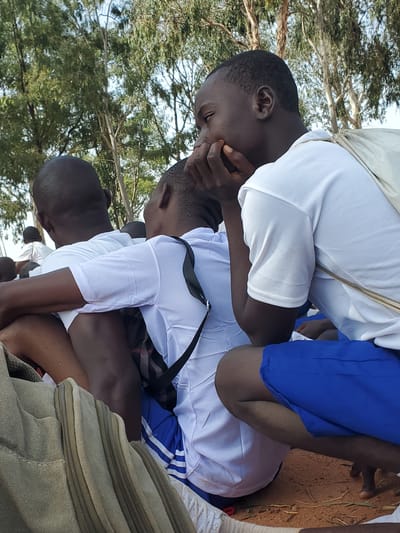 In Barakin Rafin Gora, Nigeria, four young males in white tops and blue shorts are seated on the ground, their backs to the camera, with trees and foliage in the background. The central figure, in the foreground and to the right, is leaning forward, resting his head on his hands, with his face obscured from view. To his left, another male is also leaning forward, his head and arms positioned similarly. Further left, a third individual is seated, facing away. A fourth person, visible in the distance on the left, is also seated. The ground is covered in dry earth and sparse vegetation. A canvas bag is visible in the lower left of the frame.