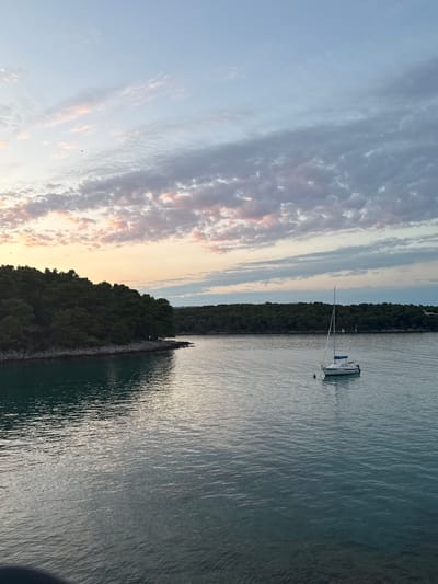A sailboat is anchored in the tranquil waters of the Adriatic Sea near Jelsa, Croatia, at dusk. The sky above is a canvas of soft blues, pinks, and grays, with wispy clouds stretching across the horizon. The sea reflects the muted colors of the sky, creating a serene and peaceful atmosphere. The shoreline is lined with dense green trees and rocky outcrops, hinting at the natural beauty of the island. The sailboat, a white vessel with a blue canopy, rests gently on the water, its mast reaching towards the sky. A single mooring buoy is visible near the boat. The overall scene evokes a sense of calm and natural splendor, characteristic of the Croatian coast.