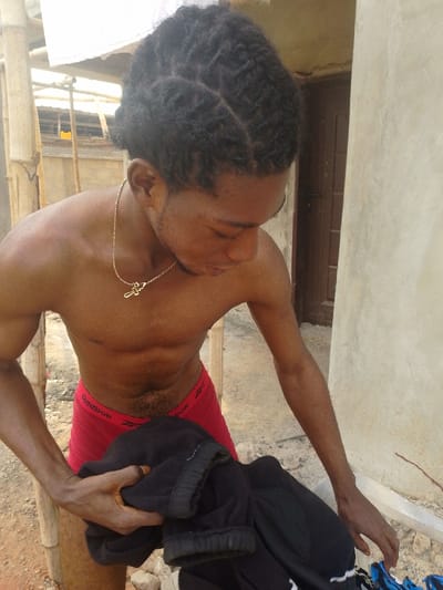 The image depicts a young, dark-skinned male situated outdoors in Enugwu-Ukwu, Nigeria. He is shirtless, revealing a muscular physique, and wears red Reebok brand boxer briefs. His dark hair is styled in intricate braids. Around his neck hangs a thin gold-colored chain with a small, stylized pendant. The subject is looking downwards, with his right hand holding a crumpled black garment, possibly sweatpants or a jacket. His left hand is extended towards the lower right, near some light-colored debris on the ground. The background features a light-colored plastered wall on the right with a dark brown door inset. To the left, bamboo poles suggest scaffolding or a temporary structure, with a white sheet or tarp draped overhead. The ground directly beneath him appears to be dirt or uneven terrain mixed with small stones.