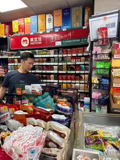 The image shows a man in a dark t-shirt standing in a convenience store. He is looking towards shelves stocked with various products. In the foreground, there are several packaged snacks, some with Chinese writing. Behind the man, shelves are filled with rows of packaged goods, including what appear to be cigarettes and boxed beverages. A large red banner with Chinese text is visible above the shelves. The store appears to be well-lit and organized. A white certificate with Chinese writing is displayed on the wall to the right, above a rack of small items. A freezer unit is visible in the lower right corner, containing packaged ice cream. The store is located in Liwan District, China.