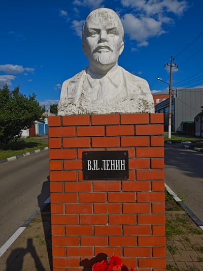 The image shows a bust of Vladimir Lenin on a brick pedestal in Novyy Oskol, Russia. The bust is made of white stone and appears weathered. It is mounted on a red brick pillar with a black plaque inscribed with "В.И. ЛЕНИН" in white Cyrillic letters. Red artificial flowers are placed at the base of the pedestal. The background shows a paved road, trees, buildings, and a blue sky with clouds. A utility pole with wires is visible on the right side of the image. The perspective suggests the photo was taken from a low angle, with the photographer's shadow visible on the left side of the road.