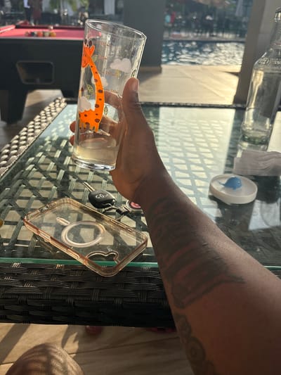 The image depicts an arm with a tattoo reaching for a glass with a giraffe design on a glass-topped wicker table. The table also holds car keys, a clear phone case, a small dish with a blue toy inside, and a glass bottle. The scene appears to be outdoors. The city of Rubuchi, Nigeria, is not evident from the image itself.