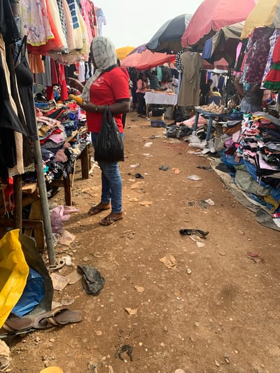 This is a photograph of an outdoor market in Nyanya, Nigeria. The ground is dirt and covered with debris, including plastic bags, paper, and other refuse. In the foreground, a person wearing a red shirt and blue jeans is standing in front of a stall displaying various clothing items. The person is holding a cob of corn and carrying a black plastic bag. The market appears to be selling used clothing, as indicated by the piles of garments on display and on the ground. Several other people can be seen in the background, as well as market stalls and umbrellas providing shade. The overall scene depicts a bustling, somewhat disorganized, marketplace environment.