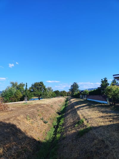 The image depicts a sunny day in Offenburg, Germany, likely near a park or recreational area. In the foreground, a dry, grassy ditch runs diagonally across the frame, casting long shadows from unseen trees. The ground on either side of the ditch is covered in dry grass and scattered leaves.
Further in the background, a paved path runs parallel to the ditch. Along the path, there are several trees with lush green foliage. A body of blue water is visible behind the trees, possibly a river or lake. A few indistinct figures can be seen on or near the path.
To the right of the image, a modern building with a dark roof and white facade is visible, bordered by a brick wall and a fence. Evergreen trees and bushes line the property. The sky is a clear, vibrant blue with a few small, wispy clouds.