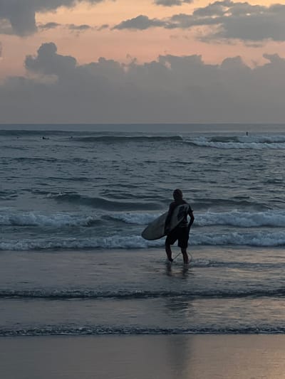 A surfer is seen waist-deep in the ocean at Canggu, Indonesia, carrying a surfboard towards the incoming waves. The scene captures a cloudy sky.