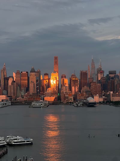 The image captures a panoramic view of the New York City skyline at sunset, as seen from Weehawken, New Jersey, United States of America. The sky is overcast with dramatic clouds, and the setting sun casts a warm, golden glow on the skyscrapers, creating a striking contrast against the darker sky. The water in the foreground reflects the colorful light of the sunset.
The skyline is dominated by numerous tall buildings, many of which are illuminated. One particularly prominent building in the center of the frame is bathed in intense orange light, its glass facade reflecting the sun's rays. Other buildings display a range of architectural styles, from modern glass towers to older brick structures. Several ferry boats are visible on the water in the lower portion of the image, including one labeled "NY WATERWAY." In the middle ground, there are piers and what appear to be floating platforms or docks. The overall scene is one of urban grandeur at dusk, with the natural beauty of the sunset transforming the cityscape.