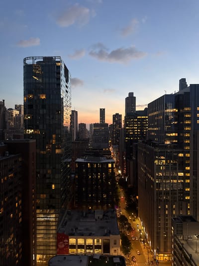 The image captures a panoramic view of the Chicago skyline at dusk. Tall buildings dominate the scene, with some illuminated from within, casting warm light against the fading blue and orange sky. The street level is visible in the distance, with car headlights and streetlights creating streaks of light. The city appears to be in motion, with lights from cars and buildings suggesting a lively urban environment. The overall impression is one of a vibrant city at the cusp of night.