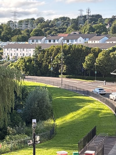 The image depicts a street scene in Cork, Ireland, with residential buildings and overhead power lines in the background. The foreground features a grassy slope with a black metal fence. A paved road runs parallel to the slope, with a silver car and a white car visible. The sky is partly cloudy, and the sun casts shadows across the grass. In the distance, tall electrical pylons are visible on a tree-covered hill. A modern apartment building with multiple windows is situated behind the trees. The scene appears to be set in a suburban area.