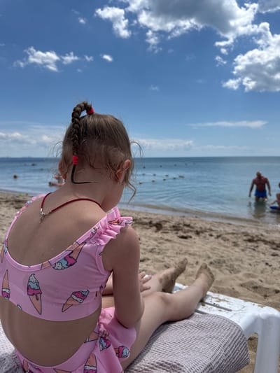 A girl in a pink ice cream-printed swimsuit is lying on a beach chair in Yevpatoriya, Ukraine. The camera is positioned behind her, showing her back and the back of her head. She has a braided pigtail with a red hair tie. In the background, there is a sandy beach, the sea with buoys, and people swimming. The sky is blue with scattered clouds. The overall scene suggests a sunny day at the beach.