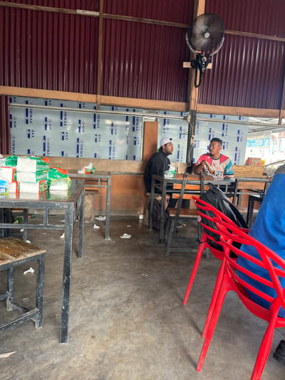 This media file shows two men sitting at tables in what appears to be an open-air establishment, possibly a restaurant or food stall, in Tambonbaroh, Indonesia. The setting features a corrugated metal wall in the background and a concrete floor. One man, wearing a colorful t-shirt, is seated on a red plastic chair and appears to be holding something in his hands, possibly food or an item he is examining. The other man, wearing a black shirt and a white cap, is seated in the foreground, facing away from the camera, with his head tilted down.
Various objects are visible, including metal tables and chairs, a large industrial fan mounted on the wall, and stacks of bagged goods, suggesting a marketplace or retail area. The overall lighting suggests daytime. The presence of banners and signs with text such as "HW MAX Germany Technology" indicates commercial branding. The scene appears casual and functional, with no strong emotional cues evident from the individuals. The overall impression is of a typical day in a local business in Indonesia.