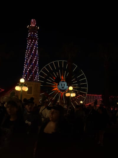 This nighttime photograph captures the vibrant scene at Disney California Adventure Park in Anaheim, USA. The iconic Mickey Mouse-themed Ferris wheel, adorned with glowing yellow lights, dominates the skyline, its smiling face illuminated. To its left, the tower of a roller coaster is visible, decorated with blue and white lights. In the foreground, blurry figures of park attendees can be seen, suggesting a bustling environment. The surrounding area is cast in darkness, with some street lamps casting a warm glow.