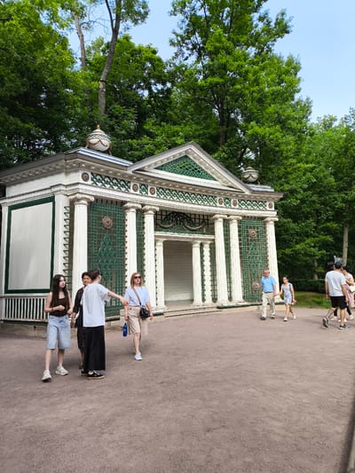 In Peterhof, Russia, a group of people stroll through the grounds of a park. In the foreground, a light brown gravel path is visible. To the left, a white pavilion with green decorative accents and classical columns stands amidst lush green trees. Four women and two men are seen walking. One woman is wearing a dark sleeveless top and denim shorts, another a white t-shirt and a long black skirt, and a third a light blue t-shirt and light-colored pants. A fourth woman, in a floral dress, stands beside another woman in a light brown t-shirt. Two men are also present, one in a blue shirt and khaki pants, and another in a gray t-shirt and black shorts. The sky above is a clear, pale blue.