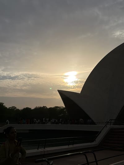 This is a vertical, silhouetted photograph of the Lotus Temple, a Baháʼí House of Worship located in Delhi, India, during sunset. The sky is filled with soft, wispy clouds, and the sun is a bright, diffused orb in the upper center, casting a warm glow and creating lens flare. The iconic, petal-like structure of the temple dominates the right side of the frame, appearing in dark silhouette against the illuminated sky. In the foreground, a woman is visible from the waist up, holding a smartphone in her left hand as if taking a picture. She is looking down at the phone and is partially silhouetted. Behind her, a railing runs horizontally. Further back, a larger group of people is gathered on a walkway or elevated platform in front of the temple, also appearing in silhouette. A line of trees forms a dark band across the horizon beneath the sky. The overall scene evokes a sense of tranquility and awe, capturing the architectural grandeur of the Lotus Temple at dusk.