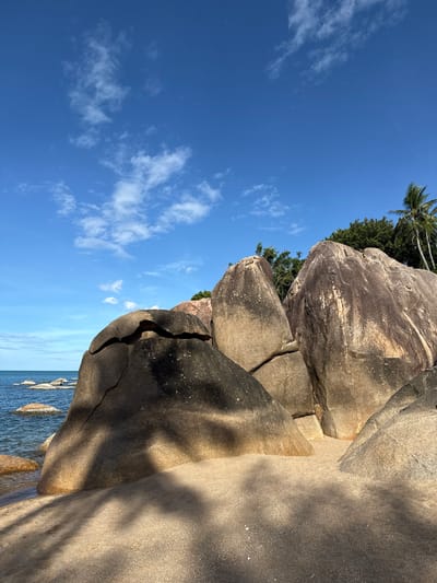 This is a landscape photograph taken at Baan Chaweng Noi, Thailand, featuring a tropical beach scene. The foreground is dominated by large, smooth, weathered granite boulders casting long shadows on the sandy beach. The sand appears fine-grained and light in color. In the middle ground, the calm, blue ocean is visible with several smaller rocks partially submerged in the water. The background shows a dense green tropical forest with palm trees, suggesting a lush environment. The sky is a clear, vibrant blue with scattered white cumulus clouds. There are no people or animals visible in the image. The overall impression is one of natural beauty and tranquility.