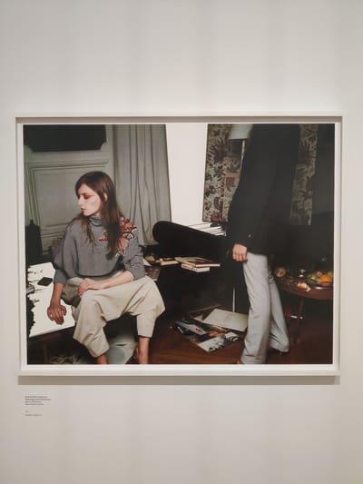 This is a photograph framed and displayed on a white wall. The photograph is oriented vertically and appears to be taken indoors. It features two individuals, a man and a woman, in what looks like a domestic setting.
The upper portion of the image shows a person from the waist down, wearing a dark coat and light-colored pants. This individual is standing near a round wooden table laden with fruit. Another person, a woman, is seated in the lower part of the image. She has long brown hair and is wearing a striped turtleneck shirt with a patterned detail on the shoulders and beige pants. Her gaze is directed downwards.
The scene is cluttered with various objects, including books and what appear to be record sleeves scattered on the floor and on furniture. The background hints at a living room or study, with patterned wallpaper and wooden flooring. The lighting suggests an indoor environment, possibly during the daytime, though no specific time of day or weather conditions are discernible.
There is text visible at the bottom right of the photograph. It reads: "Roos & Anne-Catherine- Balenciaga with M/M (Paris) Rous van Butstreten, Anne-Catiserine Lacroix) Inkjetdruk/Inkjet print". This text identifies the subjects as Roos and Anne-Catherine, mentions Balenciaga and M/M (Paris), and indicates the artwork is an inkjet print created by Rous van Butstreten and Anne-Catherine Lacroix.