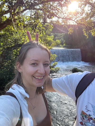 A woman is taking a selfie with a body of water and a waterfall in the background. She is smiling and has bunny ears made from her fingers on her head. She is wearing a white shirt and a brown top. The water is flowing and there are trees and sunlight in the background. There is also a bridge visible in the distance. The image appears to be taken in a natural setting, possibly in Rendang, Indonesia, given the lush vegetation and tropical feel.