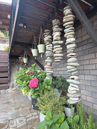The image shows a courtyard with stone and brickwork elements, and lush greenery. Hanging from the wooden overhang of a structure are several strands of stacked stones, creating decorative columns. Below these stone columns are potted plants, including hydrangeas with vibrant pink and pale yellow blooms, and a smaller green herb. To the left, a wooden staircase ascends, and a textured stone patio covers the ground. A brick wall forms the backdrop on the right side of the image. The setting appears to be a residential or public outdoor space, possibly a garden or entryway, in Nizhnyaya Shilovka, Russia. There are no people visible in the frame.