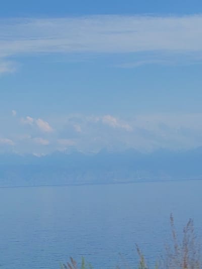 The media file depicts a wide, serene landscape under a clear blue sky dotted with wispy clouds. The scene is dominated by a vast expanse of calm water, reflecting the sky and distant mountains. The water occupies the lower half of the image, with gentle ripples suggesting a peaceful atmosphere.

In the background, a range of mountains is visible, appearing softened by distance and atmospheric haze. A thin, horizontal line stretches across the water in the mid-ground, which could be a bridge or a distant landmass. The sky takes up the upper half of the image, a soft gradient of blue with scattered, delicate clouds.

The lighting suggests it is daytime, likely mid-morning or afternoon, with soft, diffused light. The weather appears fair and calm. The overall mood is tranquil and expansive, evoking a sense of peace and natural beauty. There are no discernible people or specific objects in the foreground or mid-ground, other than the indistinct line across the water. In the bottom right corner, there are hints of dry grass or reeds, suggesting a terrestrial vantage point overlooking the water. No text is visible in the image. The landscape conveys a feeling of remoteness and natural grandeur.