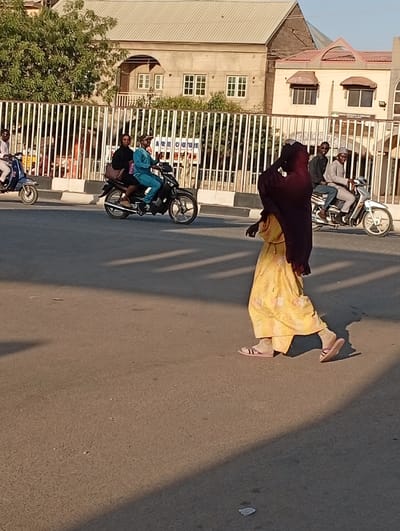 The image captures a sunlit street scene in Kabuga, Nigeria, likely during the early morning or late afternoon, as indicated by the long shadows stretching across the asphalt road.

In the foreground, a woman walks from right to left with her back mostly to the viewer. She is wearing a yellow, patterned flowing dress or long garment, a dark head covering, and light-colored sandals over socks. Her form is cast in shadow, emphasizing the light on the road.

Behind her, on the road, several motorcycles are in motion. To the left, a man in a blue outfit is driving a dark motorcycle with a female passenger behind him, who is wearing a dark top and skirt and carrying a bag. Further left, another man is riding a light-colored scooter. To the right, another man in a light-colored outfit and cap is driving a white motorcycle, with a passenger seated behind him. The driver appears to be smiling and looking towards the camera.

A white vertical-barred metal fence runs along the side of the road, separating it from an area with buildings and trees. In the background, behind the fence, are several buildings with light-colored walls and distinct rooflines, suggesting residential or commercial structures. Green foliage from trees is visible above and behind the fence.

Visible text on a sign or building behind the fence, somewhat obscured by the fence and a tree, includes parts of words like "ARBOQUE" and possibly "COLLEGE" or "OFFICE". The scene depicts a typical daily activity of commuting or movement on a street, characterized by the use of motorcycles as a common mode of transport.