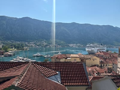 The image captures a panoramic view of Kotor, Montenegro, on a bright, sunny day. In the foreground, a cluster of red-tiled rooftops dominates the scene, some showing signs of wear. Beyond the rooftops, a bay is filled with numerous sailboats and yachts, their masts creating a delicate network against the turquoise water. Further out in the bay, a large white cruise ship is docked. The city of Kotor itself is visible along the coastline, with buildings nestled at the base of steep, verdant mountains that form a dramatic backdrop. A striking beam of light, possibly a sun pillar or lens flare, cuts vertically through the sky, intersecting the mountain range. The sky is a clear, vibrant blue, with a few wisps of clouds. There are no people visible in the image.