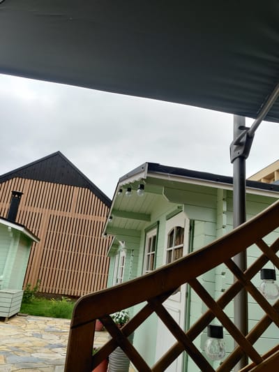 The image depicts an outdoor scene featuring two light green wooden cabins and a larger building with a distinctive slatted wooden facade, all under a cloudy sky. In the foreground, a dark wooden lattice railing runs diagonally across the frame, with two clear glass solar-powered lights attached. Beyond the railing, one of the light green cabins is partially visible, featuring white-trimmed windows and a pitched roof. Further back, another light green cabin with a similar architectural style is visible. The larger building behind the cabins has a steeply pitched dark roof and its facade is covered in vertical wooden slats, with a black chimney visible on its left side. The ground is paved with irregular, flat stones. The overall impression is of a residential or recreational property. This image was taken in Haapsalu, Estonia.