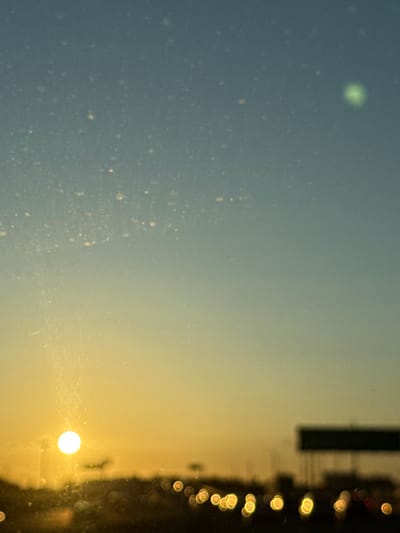 The image shows a view through a window in Oakland, United States of America. The left side of the image is a hazy blue, possibly the sky or a reflective surface. The right side transitions to yellow, with what appears to be a blurry cityscape along the edge, including a bright, circular light source, likely the sun. The blurred orbs suggest the presence of streetlights or car headlights, possibly a street at dusk or night. A tall, dark, rectangular object is also visible, possibly a building.