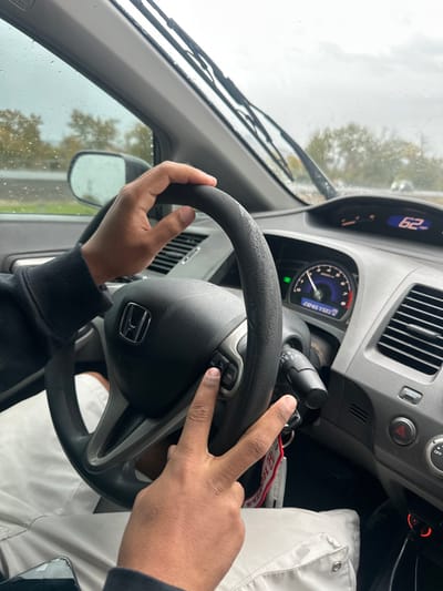 The scene is identified as the interior of a Honda vehicle, asserted to be located within Medford, United States, during rain-affected conditions. Two hands of an individual with dark skin are visible; the left hand grips the upper portion of the black steering wheel, while the right hand rests on the steering wheel's left-side controls. The right index finger is depressing a button, and the right middle finger is extended in a 'peace sign' gesture. The dashboard's digital speedometer reads '62 MPH,' and the analog tachometer displays an odometer reading of 230,456 miles. The windshield is wet with raindrops, and the wipers are in an active position. Through the windshield, an overcast sky, blurred trees, and a roadway are visible. A red Honda keychain is present near the ignition, and a dark-colored smartphone with a cracked screen is partially visible in the lower left of the frame.