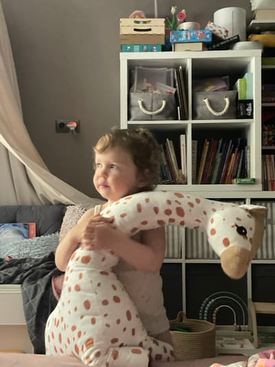 The image shows a young girl holding a large giraffe stuffed animal in her arms, standing in a room in Moscow, Russia, at the coordinates 55.659 latitude and 37.591 longitude. The girl is wearing a white tank top. The giraffe is white with brown spots. The background shows a white shelving unit filled with books, storage baskets, and other items. The room has a white curtain, a black couch with a white blanket, and a wooden step stool. The girl is looking to her right, out of frame, and is smiling. The image is taken at a slightly low angle, giving the viewer a perspective from the girl's level. The overall scene is one of playfulness and domesticity.
