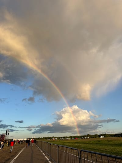 In the city of Bolgar, Russia, an outdoor event is taking place under a dramatic sky. A prominent double rainbow arches across the sky, with one arc appearing brighter and more defined than the other. The sky is a mix of bright blue patches and large, dramatic clouds that are illuminated by sunlight, giving them a golden hue in some areas and appearing gray and shadowed in others.

In the foreground, a paved path runs diagonally from the bottom left to the middle right of the image, bordered by a dark metal fence on the right side. People are walking along this path, some in small groups and others individually. They are dressed in casual clothing, with some wearing jackets.

To the left of the path, there is an open, grassy area where more people are gathered, and a large stage structure with lights and speakers is visible. Tents are also set up in this area.

Further into the background, across a grassy field, a line of parked vehicles, including cars and vans, is visible. The horizon is marked by a line of trees, and in the distance, there are some structures that appear to be communication towers. The overall impression is of a large outdoor gathering, possibly a festival or concert, occurring during or after a rainfall, as indicated by the presence of the rainbow.
