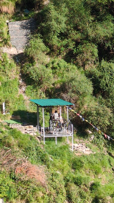 The image depicts a verdant, mountainous landscape in Bhagsu, India, under clear daylight conditions. A prominent feature is an elevated, open-sided platform or shed with a corrugated green metal roof, supported by white posts. Three individuals are present on this platform. One person, wearing a plaid shirt, stands with their back to the camera, positioned near two bicycles. Another individual is partially visible to the left of the person in the plaid shirt, and a third person is partially obscured further within the platform. Two bicycles, appearing to be mountain bikes, are securely attached to the platform structure. Multicolored prayer flags are strung diagonally from the right side of the green-roofed platform, extending downwards into the dense foliage. To the left of the platform, a flat area covered with artificial green turf is visible, alongside a vertical white cylindrical object. Stone steps and paths are integrated into the hillside, leading to and from the platform area. The surrounding environment is characterized by steep slopes covered in dense green vegetation, with some exposed earth and rock formations, including a stone retaining wall visible in the upper left portion of the image.