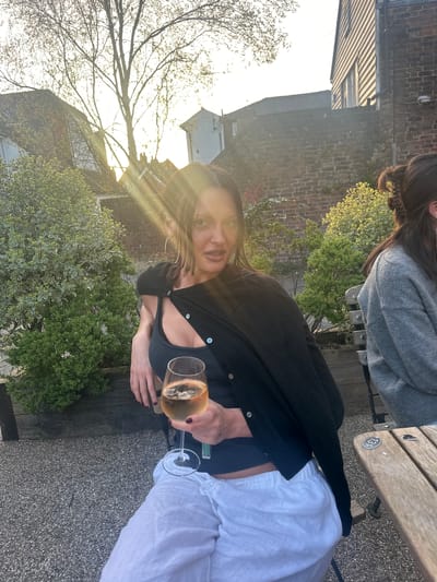Here is a description of the image:
A woman is sitting on a bench outside, holding a glass of white wine. She is wearing a black cardigan, a blue top, and white pants. Another person is partially visible on the right side of the image. The background features foliage, buildings, and the sun shining through the trees. The image appears to be taken in Tunbridge Wells, United Kingdom.
