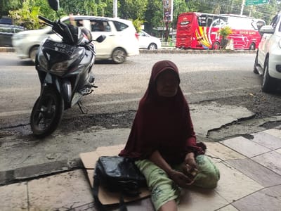 The image depicts an outdoor street scene in Situbondo, Indonesia, taken during the daytime. The primary subject is an older woman, wearing a red headscarf (hijab) and light green patterned pants, sitting on a cracked tiled sidewalk. Her face is visible, looking towards the viewer, with a somewhat neutral or somber expression. Next to her on the sidewalk is a black bag.

The woman is positioned next to a paved road with visible cracks and wear. On the road, a black scooter is parked in the foreground, facing right, with its front wheel angled. Its license plate clearly displays "P 2093 F" and "10 - 27", indicating its registration details. In the background, there is ongoing traffic, including several white cars and a distinct red public transport vehicle, likely an "angkot," which features yellow accents and text. The text on the red vehicle reads "SELAMAT DATANG DI SITUBONDO" (Welcome to Situbondo) and "GENTONG BIRU." Further in the background, trees and other buildings can be vaguely seen, suggesting an urban or semi-urban setting. The overall scene conveys a candid moment of street life.