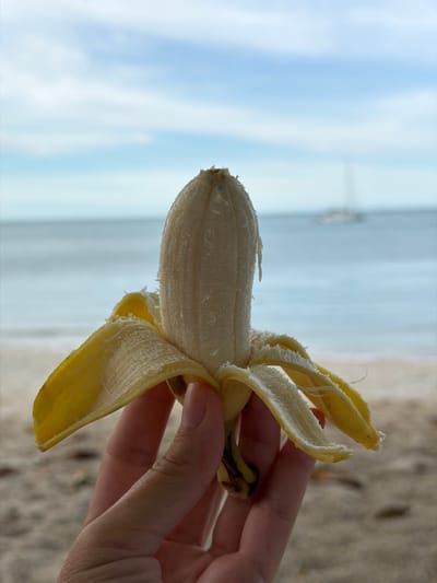 A hand holds a partially peeled banana with the peel splayed out like a flower. The banana is light yellow and smooth, with some white stringy bits hanging from it. The hand is pale with short fingernails. In the background, the ocean meets a sandy beach under a pale blue sky with some clouds. A faint outline of a boat can be seen on the water. This scene appears to be set in Baan Plai Laem, Thailand.